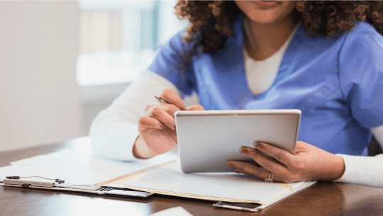 A person wearing blue scrubs uses a tablet while sitting at a table with paperwork and a tablet.