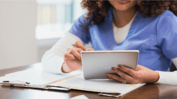 A person wearing blue scrubs uses a tablet while sitting at a table with paperwork and a tablet.