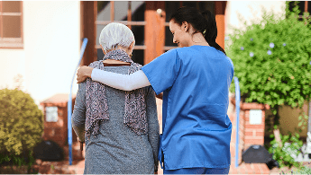 Healthcare professional with her arm around an elderly patient, walking up to a building image