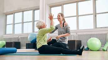 Elderly woman sat down on a mat in a fitness studio, high-fiving a fitness instructor image