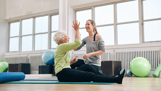 Elderly woman sat down on a mat in a fitness studio, high-fiving a fitness instructor image