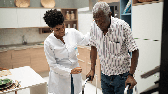 Elderly man holding himself up with a walking frame, stood alongside a healthcare professional image