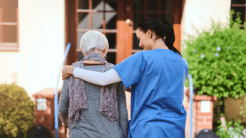 A healthcare worker in blue scrubs walks outside with an elderly woman, supporting her with an arm around her shoulders.
