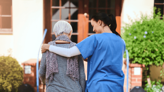 A healthcare worker in blue scrubs walks outside with an elderly woman, supporting her with an arm around her shoulders.