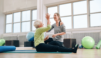 Elderly woman sat down on a mat in a fitness studio, high-fiving a fitness instructor image