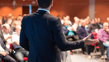A man stood at the front of a lecture hall giving a talk to a room full of people image