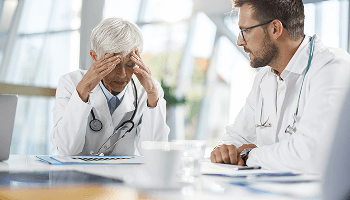 Healthcare professional at a table with her head in her hands, sat next to a colleague image