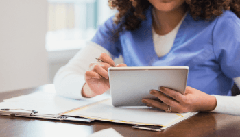 A person wearing blue scrubs uses a tablet while sitting at a table with paperwork and a tablet