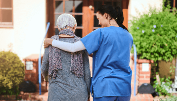 Healthcare professional with her arm around an elderly patient, walking up to a building image