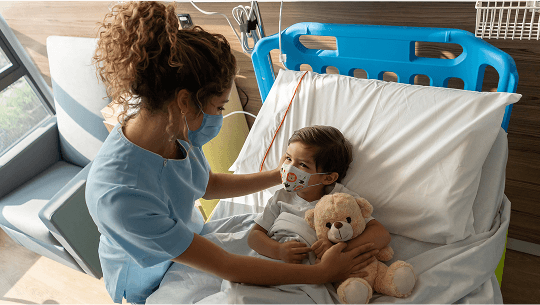 Young child holding a teddy bear in a hospital bed whilst a healthcare professional provides care image