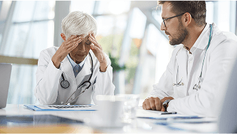 Healthcare professional at a table with her head in her hands, sat next to a colleague image