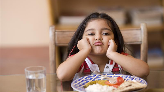 A young girl sits at a table with her head in her hands, looking bored or unhappy, in front of a plate of food and a glass of water.