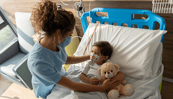 Young child holding a teddy bear in a hospital bed whilst a healthcare professional provides care image